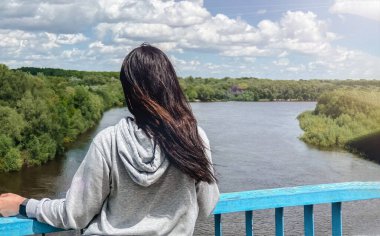 Koyu saçlı, beyaz bir kız sırtı açık gri bir ceketle duruyor ve nehre ve ormana doğru bakıyor.