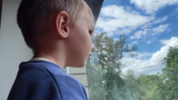 Menino caucasiano olha pela janela e sorri para a câmera no verão em uma casa particular — Vídeo de Stock