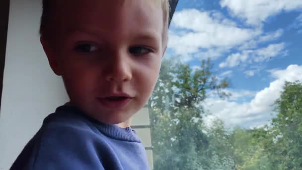 Little caucasian boy looks out the window and then smiles at the camera in the summer in a private house — Stock Video