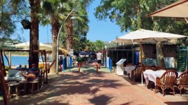 Türkiye 'de bir tatil köyü boyunca palmiye ağaçları, dükkanlar, plaj şemsiyeleri arasında mavi gökyüzüne karşı yürüyün