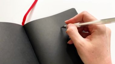 Beyaz mürekkepli yazıtlar için siyah kağıtlı bir defterin yakın plan gösterimi. Bir kadının eli beyaz mürekkeple bir kalp çizer.