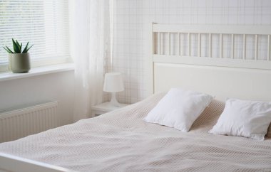 Odanın içi beyaz tonlarda, yatak, komodin, masa lambası ve pencere eşiğinde bir çiçek.