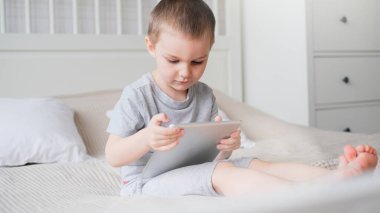 A small caucasian boy in a light room in a gray T-shirt plays with a tablet while sitting on the couch. 