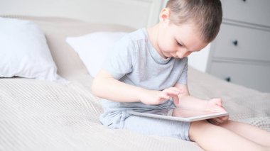 Aydınlık bir odada gri tişörtlü beyaz bir çocuk kanepede otururken tabletle oynuyor.. 