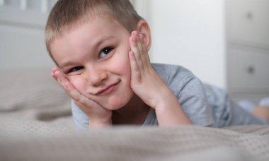 Gri tişörtlü, küçük, mutlu, beyaz bir çocuğu yakından çek ve yatağa uzan ve gülümse..