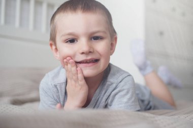 Gri tişörtlü, küçük, mutlu, beyaz bir çocuğu yakından çek ve yatağa uzan ve gülümse..