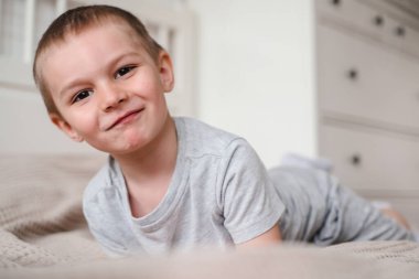 Gri tişörtlü, küçük, mutlu, beyaz bir çocuğu yakından çek ve yatağa uzan ve gülümse..