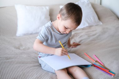 A small caucasian boy in a gray T-shirt and shorts on the bed draws with colored pencils in a sketchbook. Childrens creativity and development.