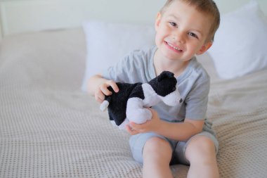 Close-up of a small happy caucasian boy in a gray t-shirt lies on the bed with a toy dog.
