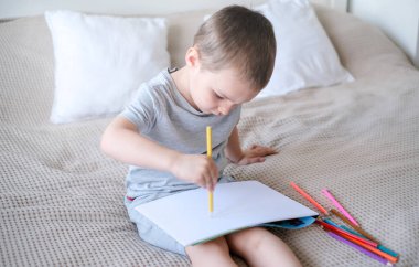 A small caucasian boy in a gray T-shirt and shorts on the bed draws with colored pencils in a sketchbook. Childrens creativity and development.