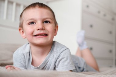 Gri tişörtlü, küçük, mutlu, beyaz bir çocuğu yakından çek ve yatağa uzan ve gülümse..
