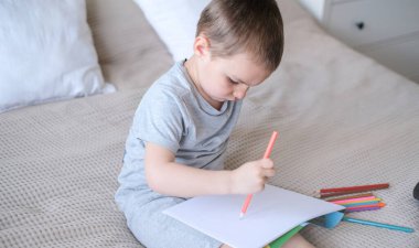 A small caucasian boy in a gray T-shirt and shorts on the bed draws with colored pencils in a sketchbook. Childrens creativity and development.