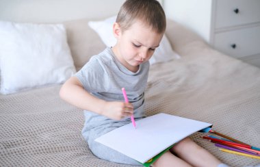 A small caucasian boy in a gray T-shirt and shorts on the bed draws with colored pencils in a sketchbook. Childrens creativity and development.