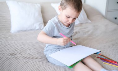 A small caucasian boy in a gray T-shirt and shorts on the bed draws with colored pencils in a sketchbook. Childrens creativity and development.