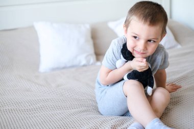 Close-up of a small happy caucasian boy in a gray t-shirt lies on the bed with a toy dog.