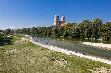 Münih, Almanya - 26 Eylül 2016. St. Maximilian 'ın kilisesi ağaçların arkasından gözetliyor. İnsanlar güneşli bir günde isar nehrinin kıyısında geziniyor, oynuyor ve konuşuyor..