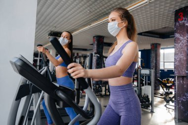 Salgın sırasında maske takan iki güzel kız spor salonunda egzersiz yapıyor. Halka açık yerlerde sosyal mesafeler.