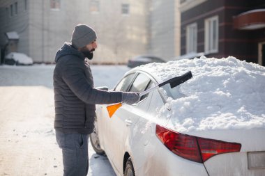 Genç bir adam güneşli, soğuk bir günde kar yağdıktan sonra arabasını temizler..