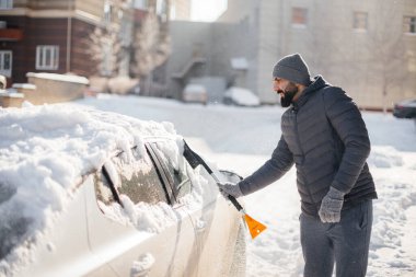 Genç bir adam güneşli, soğuk bir günde kar yağdıktan sonra arabasını temizler..