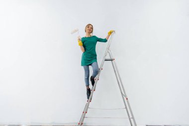 Merdivendeki genç bir kız beyaz bir duvarı silindirle boyuyor. İç kısmın tamiri.