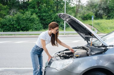 Genç bir kız otoyolun ortasında bozuk bir arabanın yanında duruyor ve kaputun altına bakıyor. Aracın bozulması ve arızası. Yardım bekliyorum..