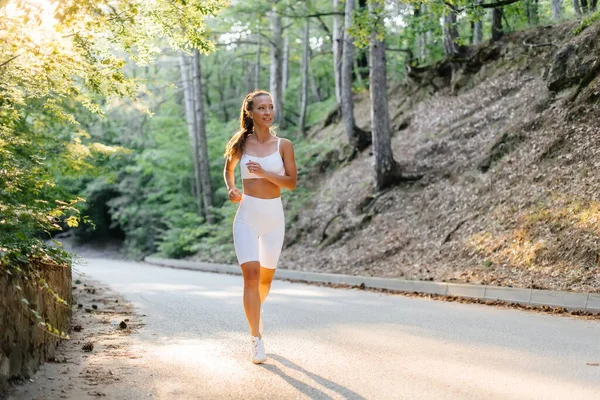 白いスポーツ服を着た若い美しい少女が 日没の間 鬱蒼とした森の中の道路上で走っています 新鮮な空気の中でスポーツをしている 健康的なライフスタイル — ストック写真