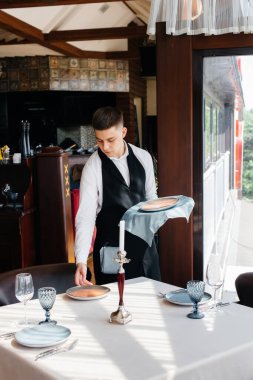 Şık üniformalı genç bir erkek garson güzel bir gurme restoranında masaya servis yapmakla meşgul. Restoran faaliyetleri, en üst düzeyde..