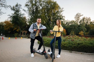 Genç ve güzel bir çift sıcak bir sonbahar gününde parkta elektrikli scooter kullanıyor. Hobiler ve eğlence..