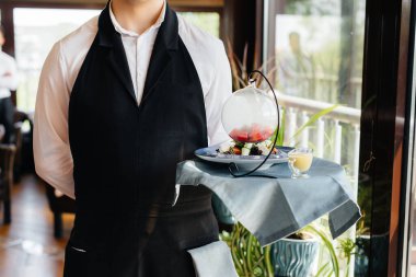 Şık bir üniforma içindeki genç bir garson, güzel bir restoranın yakın çekiminde masanın yanındaki tepside nefis bir tabakla duruyor. Restoran faaliyetleri, en üst düzeyde..