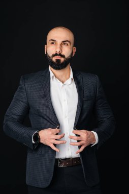 Beyaz gömlekli ve ceketli genç sakallı bir adam siyah arka planda duruyor..