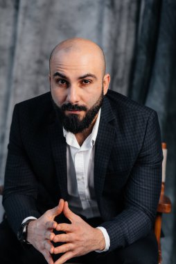 Beyaz gömlekli ve takım elbiseli genç sakallı bir adam stüdyoda bir sandalyeye oturur..