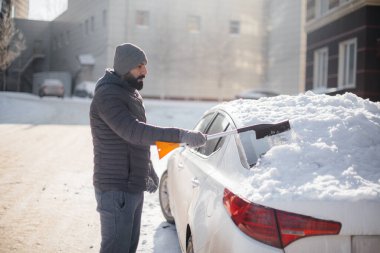 Genç bir adam güneşli, soğuk bir günde kar yağdıktan sonra arabasını temizler..