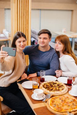 Bir grup neşeli genç arkadaş bir kafede oturup konuşuyorlar ve telefonda selfie çekiyorlar. Pizzacıda öğle yemeği..