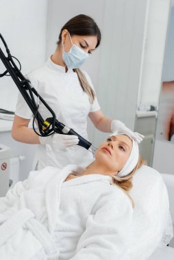 Genç bir kız güzellik salonunda yüzünün derisi için kömür soyuyordu. Lazer darbeleri deriyi temizler. Donanım kozmetolojisi. Fototermoliz süreci, deriyi ısıtır..