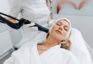 Genç bir kız güzellik salonunda yüzünün derisi için kömür soyuyordu. Lazer darbeleri deriyi temizler. Donanım kozmetolojisi. Fototermoliz süreci, deriyi ısıtır..