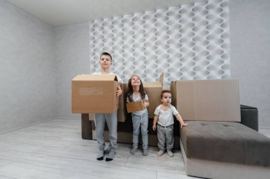 Erkek ve kız kardeşler karton kutuların ve kanepenin arka planında yeni bir daireye taşınıp eğleniyorlar. Gayrimenkul alımı. Ev partisi, teslimat..