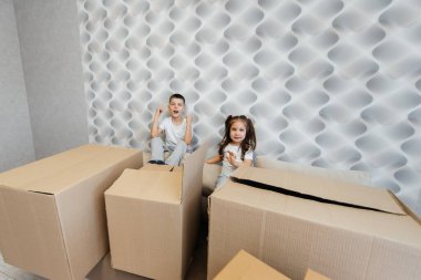 Erkek ve kız kardeş kanepedeki karton kutuların arka planına karşı yeni bir daireye taşınıp eğleniyorlar. Gayrimenkul alımı. Ev partisi, teslimat..