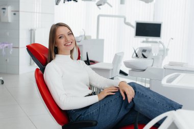 Güzel genç bir kadın modern beyaz dişçilikte kırmızı bir dişçi koltuğunda oturuyor ve gülümsüyor. Diş protezi, tedavi ve diş beyazlatma..