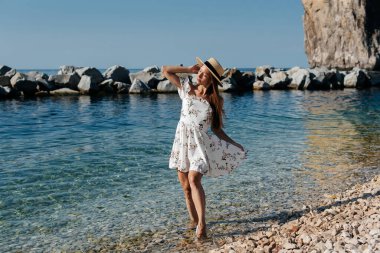 Şapkalı, gözlüklü ve açık renk elbiseli güzel bir kadın güneşli bir günde okyanus kıyısında büyük kayaların arkasında yürüyor. Turizm ve turizm gezileri.