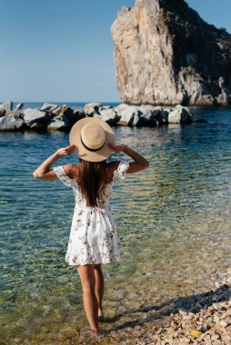 Şapkalı ve sırtında açık renk elbisesi olan güzel bir kadın güneşli bir günde okyanus kıyısında büyük kayaların arkasında yürüyor. Turizm ve tatil gezisi.