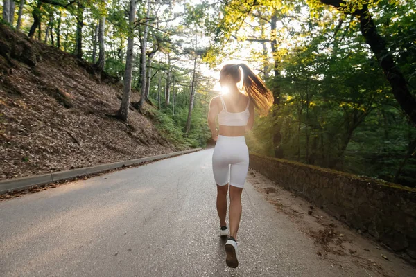 白いスポーツ服を着た若い美しい少女が 日没の間 鬱蒼とした森の中の道路で彼女と一緒に走っています 新鮮な空気の中でスポーツをしている 健康的なライフスタイル — ストック写真