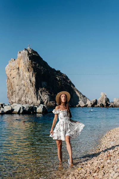 Şapkalı, gözlüklü ve açık renk elbiseli güzel bir kadın güneşli bir günde okyanus kıyısında büyük kayaların arkasında yürüyor. Turizm ve turizm gezileri.