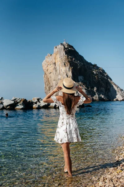 Şapkalı ve sırtında açık renk elbisesi olan güzel bir kadın güneşli bir günde okyanus kıyısında büyük kayaların arkasında yürüyor. Turizm ve tatil gezisi.