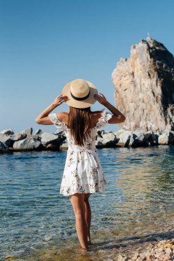 Şapkalı ve sırtında açık renk elbisesi olan güzel bir kadın güneşli bir günde okyanus kıyısında büyük kayaların arkasında yürüyor. Turizm ve tatil gezisi.
