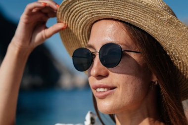 Şapkalı, gözlüklü ve açık renk elbiseli güzel bir kadın güneşli bir günde okyanus kıyısında büyük kayaların arkasında yürüyor. Turizm ve turizm gezileri.