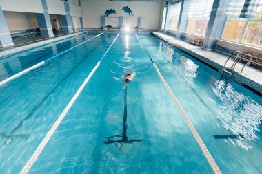 Genç bir adam modern bir yüzme havuzunda antrenman yapar ve yüzer. Spor gelişimi. Yarışmalara hazırlık ve sağlıklı bir yaşam tarzı. Su tedavileri ve sağlıklı bir yaşam tarzı..