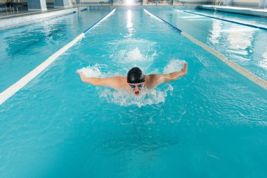 Genç bir adam modern bir yüzme havuzunda antrenman yapar ve yüzer. Spor gelişimi. Yarışmalara hazırlık ve sağlıklı bir yaşam tarzı. Su tedavileri ve sağlıklı bir yaşam tarzı..