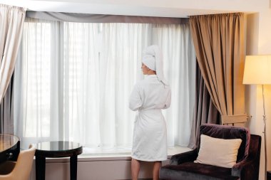 Beyaz önlüklü güzel bir kız otel odasının penceresinin yanında duruyor ve kahve içiyor. Dinlen ve seyahat et. Otel eğlencesi ve turizm..