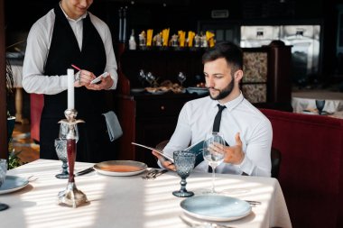 İyi bir restoranda genç bir işadamı menüyü inceler ve şık önlüklü genç bir garsona sipariş verir. Müşteri hizmetleri. Restorandaki masa servisi..