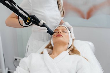 Genç bir kız güzellik salonunda yüzünün derisi için kömür soyuyordu. Lazer darbeleri deriyi temizler. Donanım kozmetolojisi. Fototermoliz süreci, deriyi ısıtır..